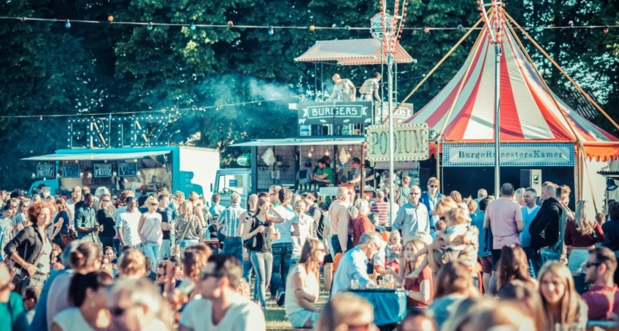 Busy festival grounds in the sun with food trucks, a stage and a red-and-white circus tent